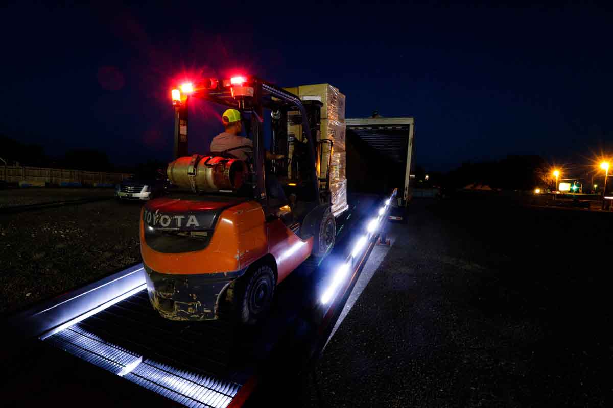 Operator using a Toyota forklift at night to unload a load from a truck via a ramp. Tail-lights alert other workers that the lift truck is in operation.